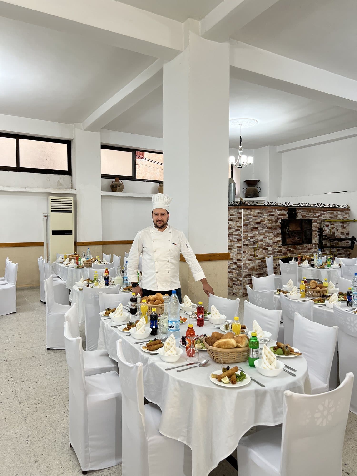 Un chef en tenue blanche dans une salle aux tables nappées de blanc, dressées pour le repas.